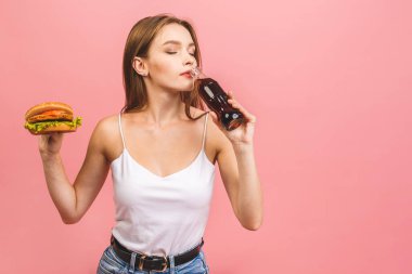 Burger yiyen genç ve aç bir kadının portresi. Pembe arka planda fast food yiyen öğrencilerin izole edilmiş portresi. Diyet kavramı. 