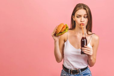 Burger yiyen genç ve aç bir kadının portresi. Pembe arka planda fast food yiyen öğrencilerin izole edilmiş portresi. Diyet kavramı. 