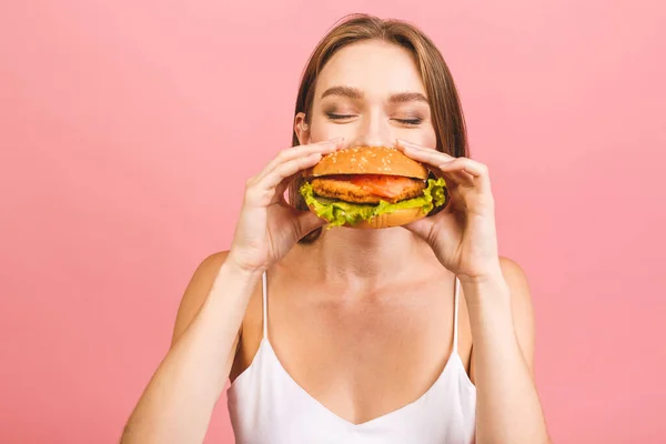 Burger yiyen genç ve aç bir kadının portresi. Pembe arka planda fast food yiyen öğrencilerin izole edilmiş portresi. Diyet kavramı. 