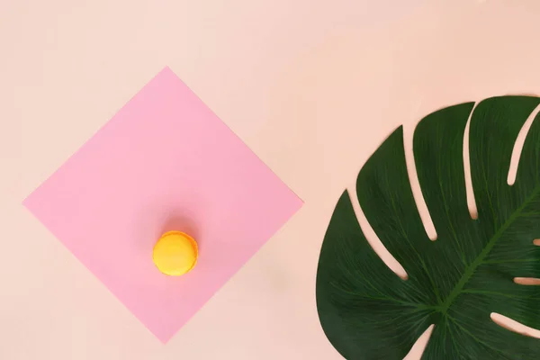 Düz kompozisyon ve pembe kağıt karede duran sarı bir badem kurabiyesi. Sağ tarafta canavarın tropikal yaprağı var. Açık pembe arkaplanda kağıt desenli nesneler. El yapımı tatlılar.