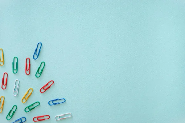 Açık mavi arkaplanda çok renkli ataçların yatay yakın çekim düz kompozisyonu. Sol alt köşede yeşil, kırmızı, sarı ve mavi renkli eşyalar var. Ofis malzemeleri konsepti