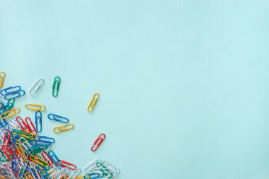 Açık mavi arkaplanda çok renkli ataçların yatay yakın çekim düz kompozisyonu. Sol alt köşede yeşil, kırmızı, sarı, beyaz ve mavi nesneler yığını var. Ofis malzemeleri