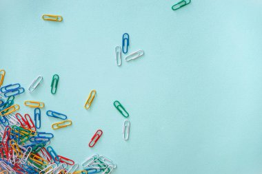 Açık mavi arkaplanda çok renkli ataçların yatay yakın çekim düz kompozisyonu. Sol alt köşede yeşil, kırmızı, sarı, beyaz ve mavi nesneler yığını var. Ataçların parçası