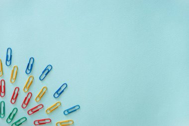 Açık mavi arkaplanda çok renkli ataçların yatay yakın çekim düz kompozisyonu. Sol alt köşede yeşil, kırmızı, sarı ve mavi renkli eşyalar var. Ofis malzemeleri konsepti