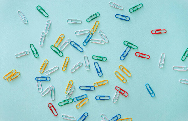 Horizontal close-up flat lay composition of multi-colored paper clips on a light blue background. Items of green, red, yellow and blue colour located randomly throughout the picture. Office supplies