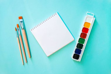 Yatay minimalistik düz kompozisyon, temiz sayfa not defteri, turuncu kazıklı üç açık bej fırça, ahşap doku ve açık mavi arkaplanda suluboya.