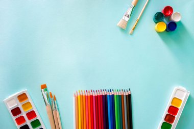 Yatay yakın çekim düz kompozisyonu, turuncu kazıklı ve ahşap desenli üç açık bej fırça, iki set suluboya boya boya, renkli kalemler ve açık mavi renkte beş gouache boya.