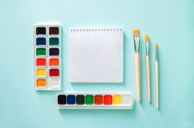 Yatay minimalistik düz kompozisyon, temiz sayfa not defteri, turuncu ve ahşap desenli üç açık bej fırça ve açık mavi üzerine iki set suluboya.