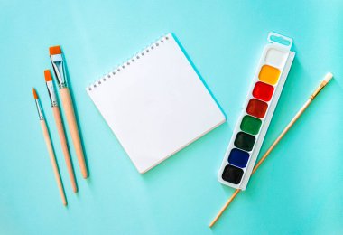 Yatay minimalistik düz kompozisyon, temiz sayfa not defteri, turuncu ve ahşap desenli üç açık bej fırça, beyaz kazıklı bir fırça ve suluboya