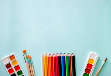 Yatay yakın çekim düz kompozisyonu, turuncu kazıklı ve ahşap desenli üç açık bej fırça, iki set suluboya boya boya, renkli kalemler ve açık mavi renkte beş gouache boya.