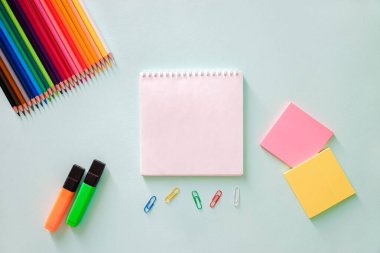 Renkli kalemler, kırtasiye etiketleri pembe ve limon rengi, ataç, turuncu ve yeşil fosforlu kalem ve açık mavi arkaplan üzerine beyaz not defteri. Yatay fotoğraf. Düz minimalist kompozisyon. Okula, üniversiteye, eğitime 