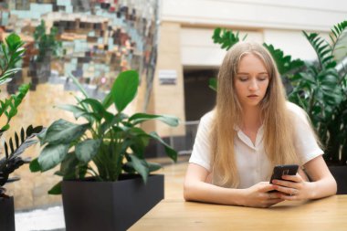 Güzel, neşeli, beyaz, sarışın bir iş kadını masanın üzerinde oturmuş, telefona bakıyor ve düşünüyor. Bir şirkette başarılı genç kadın kavramı. Bir şirkette başarılı genç kadın kavramı. Beyaz gömlekli kız.. 