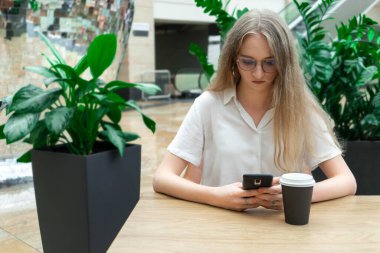 Güzel, neşeli, beyaz, sarışın bir iş kadını masanın üzerinde oturmuş, telefona bakıyor ve düşünüyor. Bir şirkette başarılı genç kadın kavramı. Beyaz gömlekli kız. Yatay fotoğraf. Bitkilerle bulanık bir arkaplan. Başla.