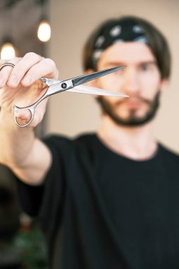 Keskin bir berber makası. Siyah tişörtlü ve bandanalı bulanık bir kuaförün yakın plan fotoğrafı. Sağ elinde makasla kameraya doğru uzanıyor..