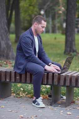 Serbest çalışan, mavi giyimli, sıradan bir süit, dizüstü bilgisayarla çalışıyor. Broker parktaki ahşap bankta oturuyor ve işini yapıyor. Aklında borsa çizelgeleri var. Serbest çalışma ve serbest çalışma kavramı.