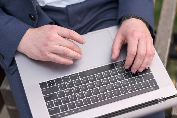 Close-up of male Hands on the keyboard with Russian letters of computer. Freelancer working out of office. Man using laptop while sitting on the bench in park. Small depth of field. Selective focus.