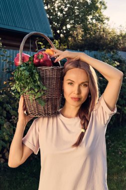Handsome white Woman wearing t-shirt holds on shoulder wicker basket with autumn harvest of bell peppers, parsley, eggplants, cucumbers and tomatoes. Concept of gardening and harvesting. clipart