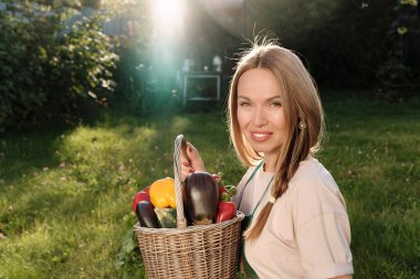 Önlük takan yakışıklı beyaz kadın elinde sepetin içinde dolma biber, maydanoz, patlıcan, salatalık ve domates hasadı ile el ele tutuşuyor. Bahçe işleri ve hasat kavramı.