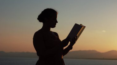 Gün batımında deniz kenarında okuyan genç bir kadının silueti.