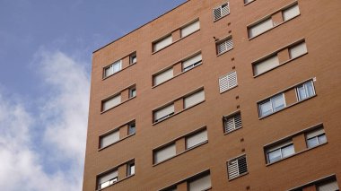 Üniformalı tuğladan dış görünüşü ve parçalı bulutlu bir gökyüzünün altında birden fazla penceresi olan yüksek bir apartmanın alçak açılı görüntüsü..