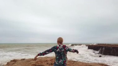 Kız, fırtına sırasında sırtı deniz kıyısında dururken ellerini kaldırır..