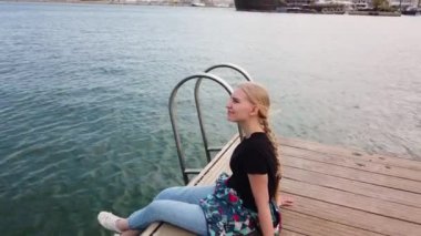 Limandaki iskele kıyısında tek başına oturan kız park etmiş yelkenlilerin önünde ve güzel manzarası olan dinleniyor..