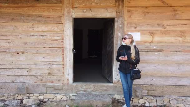 Γυναίκα που χρησιμοποιεί έξυπνο τηλέφωνο κοντά σε ένα ξύλινο σπίτι σε ένα παλιό χωριό. — Αρχείο Βίντεο
