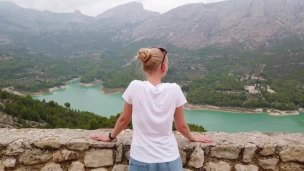 Ein junges Mädchen blickt von der Mauer eines alten Schlosses auf eine schöne Aussicht mit blauem See und Bergen — Stockvideo