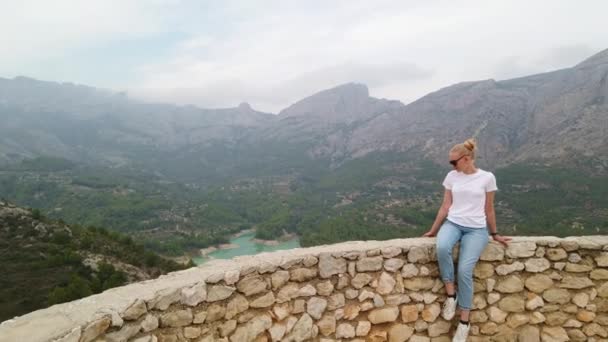 Bir kadın oturuyor ve mavi gölü ve eski bir kalenin duvarından dağları olan güzel bir manzaraya bakıyor. — Stok video