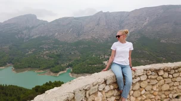 Donna seduta a guardare una splendida vista con un lago blu e montagne dal muro di un vecchio castello — Video Stock