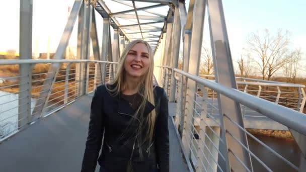 Menina loira andando na ponte e olha para a câmera ao pôr do sol . — Vídeo de Stock