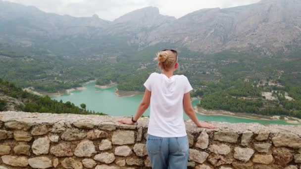 Uma jovem mulher olha para uma bela vista com um lago azul e montanhas a partir da parede de um antigo castelo — Vídeo de Stock