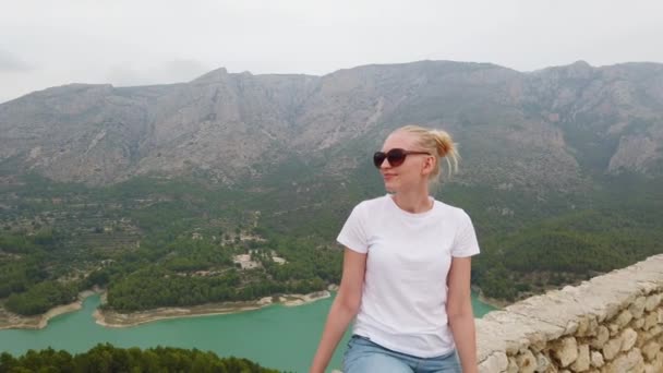 Mulher sentada e olhando para uma bela vista com um lago azul e montanhas a partir da parede de um castelo velho — Vídeo de Stock