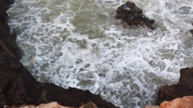 Rüzgârlı ve yağmurlu bir günde kumsalda kayalara çarpan büyük dalgalar çok fazla beyaz köpük saçıyor. Ağır çekimde. 4K