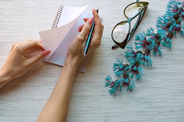 Female hands with a pen in their hands write in a notebook, on a white wooden background, near glasses, and blue cornflowers. View from above. Glasses for sight. Pink nails, blue pen.