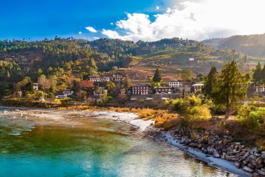 Mo Chhu Nehri güneşli bir günde, Punakha, Butan. Punakha Dzong yakınlarındaki tahta kantilever köprüsünden nehre Punakha şehrinin ve Himalaya dağlarının evlerinin manzarası.