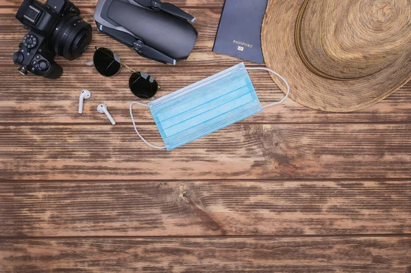 Tahta arka planda seyahat aksesuarları: yüz maskesi, bluetooth kulaklık, pasaport, hasır şapka, drone, aynasız kamera ve güneş gözlüğü. Üst görünüm noktası.