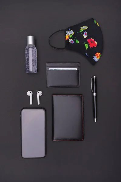 Vertical shot of travel accessories kit on black background: face mask, antibacterial gel, passport, cell phone, bluetooth headphones, ballpoint pen and card holder. Top view point.