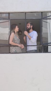 Pencerenin önünde konuşan bir çift, binanın dışından bir video. Pencere evinde gülümseyen ve rahatlatan genç çift, yavaş çekimde dikey video.