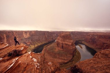 Bulutlu bir havada Colorado Nehri 'nde at nalı üzerinde fotoğraf çeken bir adam. At nalı kıvrımının panoramik görüntüsü