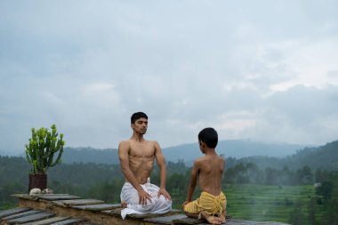 Genç yakışıklı bir çocuk, dağ sırasının ortasındaki bir evin çatısında bir çocukla yoga yapıyor. Yoga ve fitness.