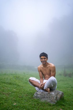 Delikanlı dhoti giyiyor, bir kayanın üzerinde oturuyor, sisli bir sabahta yeşil alanda açık..