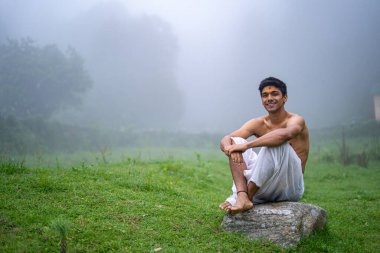 Delikanlı dhoti giyiyor, bir kayanın üzerinde oturuyor, sisli bir sabahta yeşil alanda açık..