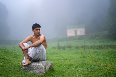 Delikanlı dhoti giyiyor, bir kayanın üzerinde oturuyor, sisli bir sabahta yeşil alanda açık..
