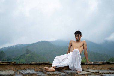 Dhoti 'li ve arka planda bulanık dağlı gözlüklü genç bir adam oturuyor..