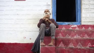 Uzun beyaz sakallı ve bıyıklı yaşlı bir adam bacak bacak üstüne atmış merdivenlerde oturuyor.