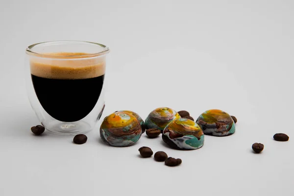 Tam siyah kremalı espresso bardağı ve etraflarında kavrulmuş kahverengi kahve çekirdekleri ve fotokopi alanı ile beyaz arka planda izole edilmiş gezegenlere benzeyen renkli yuvarlak sütlü çikolatalar.