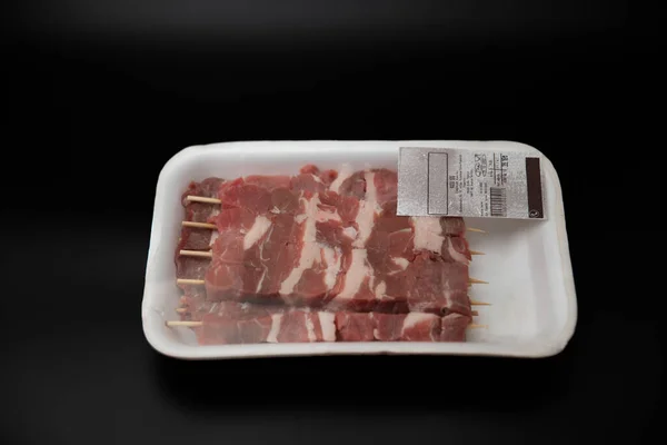 Üzerinde barkod olan beyaz bir plastik kapta tahta şişler üzerinde ham doğranmış et (küp şeklinde doğranmış), metin için metin alanı olan siyah arkaplan üzerinde izole edilmiş.