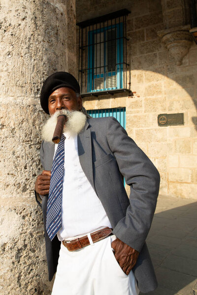 Havana / Cuba - 04.16.2015: Afro cuban old man with a long white beard wearing a suit and a black military beret, smoking a big cigar