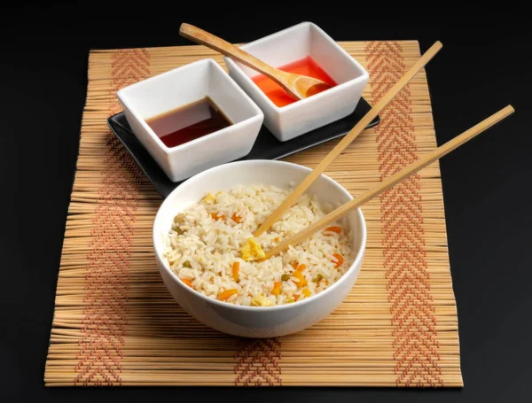 Chinese food bowl of rice with chopsticks and soy sauce and sweet and sour sauce on bamboo trivet and black background
